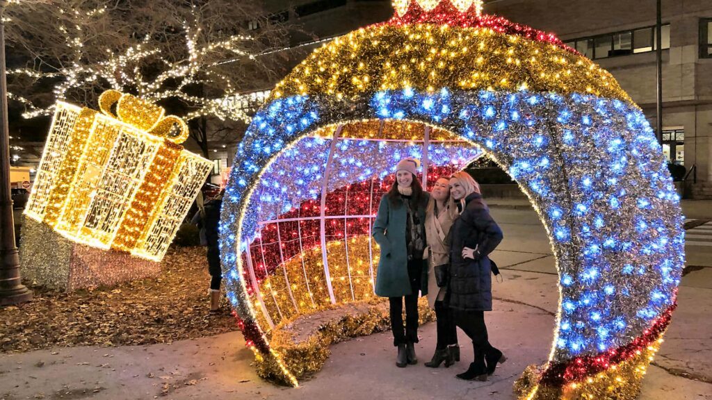 A group of 3 girls under a large lit Christmas ornament diagram