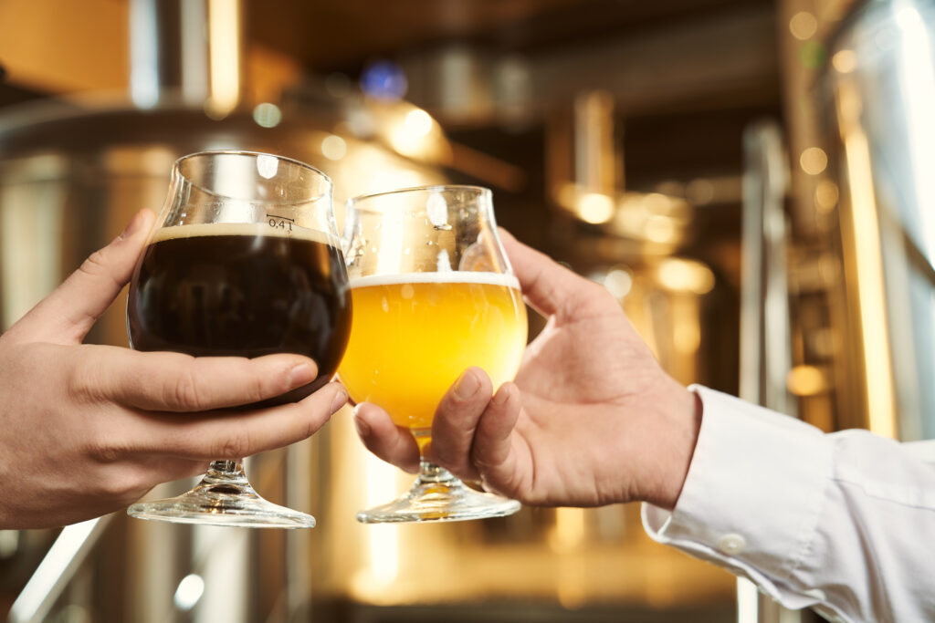 Close up of two male hands holding glasses with beer. Assortment of drinks. Dark and light beer. Industry of brewing beer. Equipment of modern mini brewery on background.